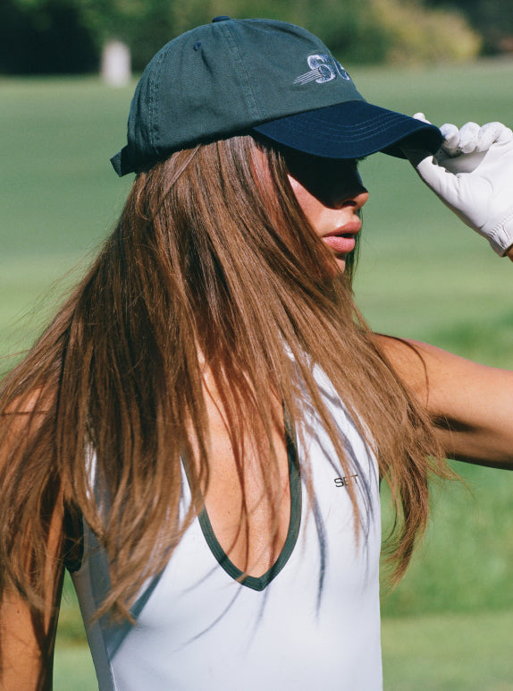 A woman on a golf course wearing a dark green and navy baseball cap with a white golf shirt and white glove, looking over her shoulder.