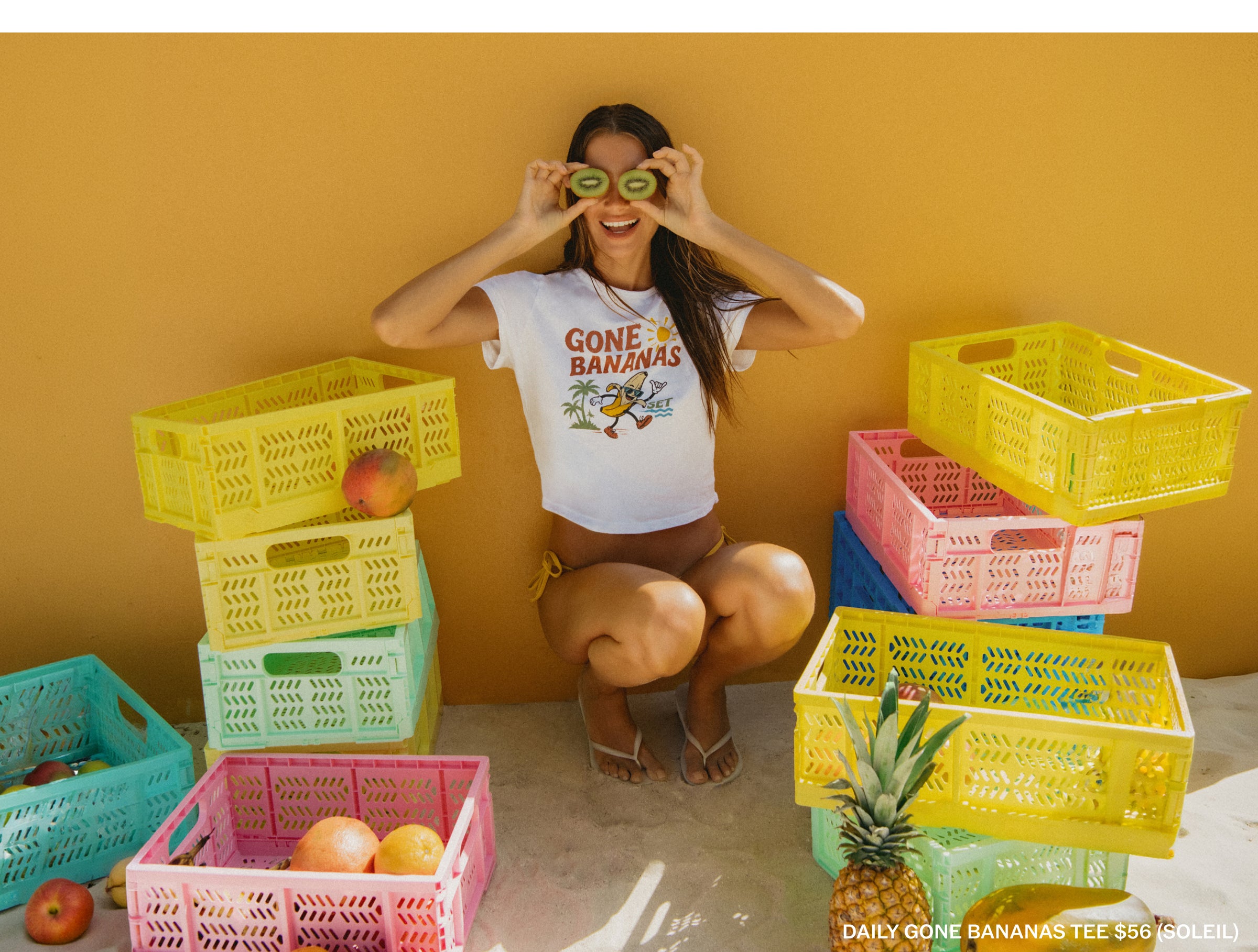 A woman crouches against a mustard yellow wall wearing a white graphic T-shirt that reads 'GONE BANANAS' with a banana illustration. She is surrounded by stacked translucent plastic crates in yellow, pink, blue, and green, with fruit props including bananas, apples, and a pineapple.