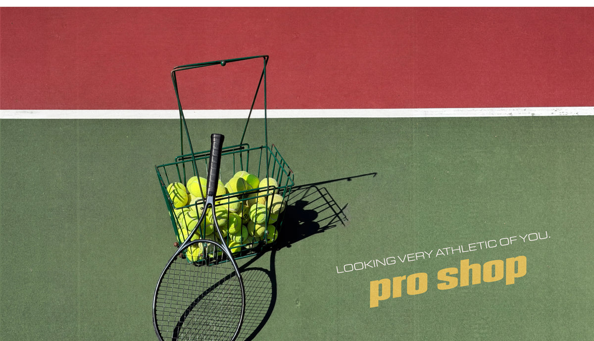 A green wire tennis ball basket filled with yellow tennis balls on a tennis court, with a tennis racket leaning in front. The background shows a red and green tennis court with the text 'LOOKING VERY ATHLETIC OF YOU.