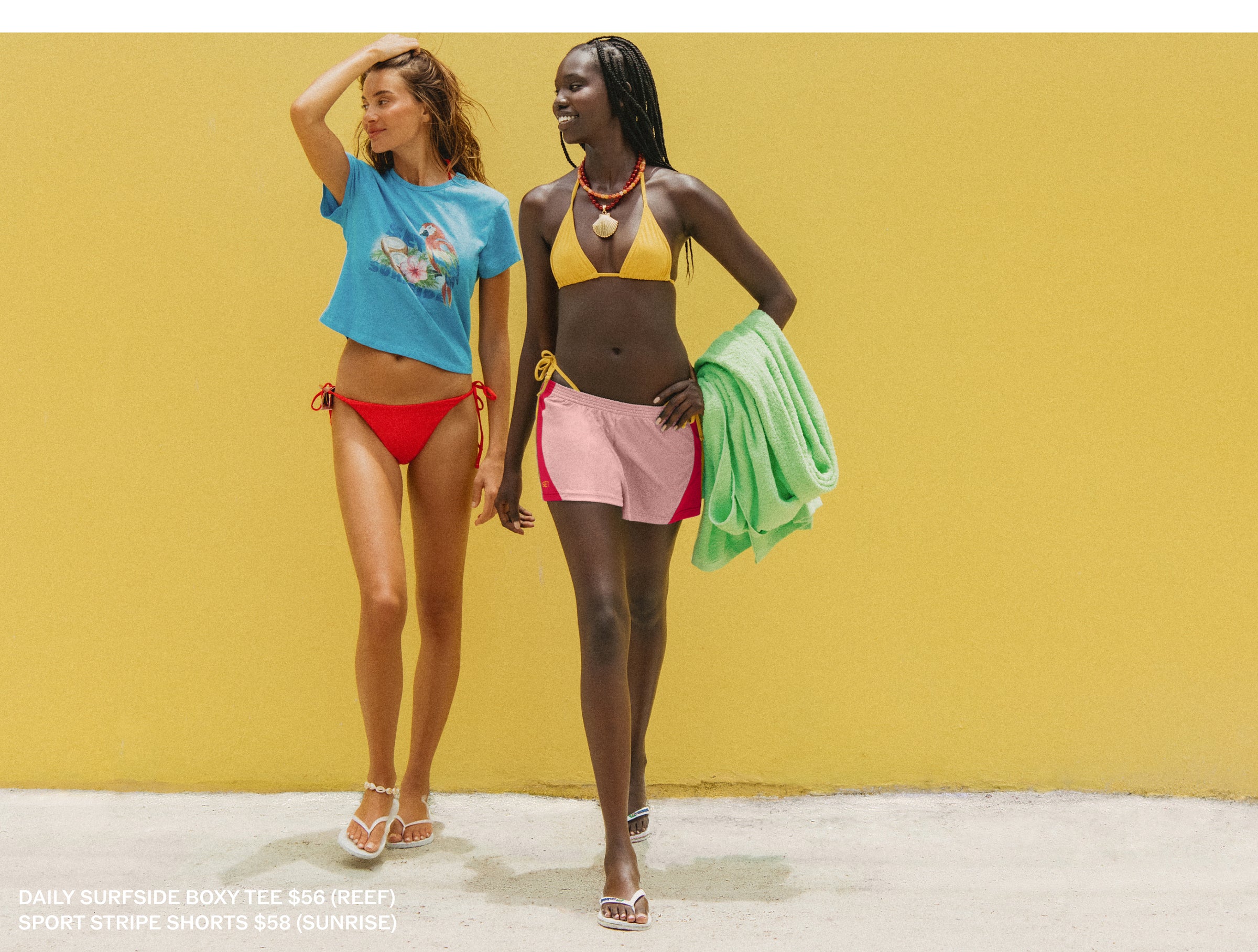 Two models walking side-by-side against a solid yellow wall, showcasing swimwear. The model on the left wears a light blue graphic t-shirt and red bikini bottoms.