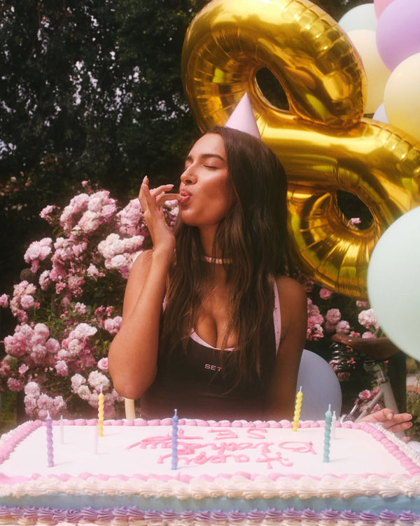Person blowing out candles on a birthday cake with balloons and flowers in the background