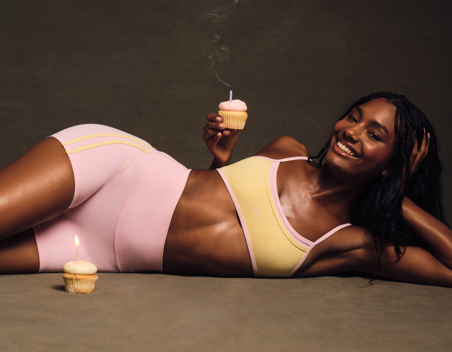Woman in pink and yellow lingerie holding a cupcake with a lit candle on a dark background