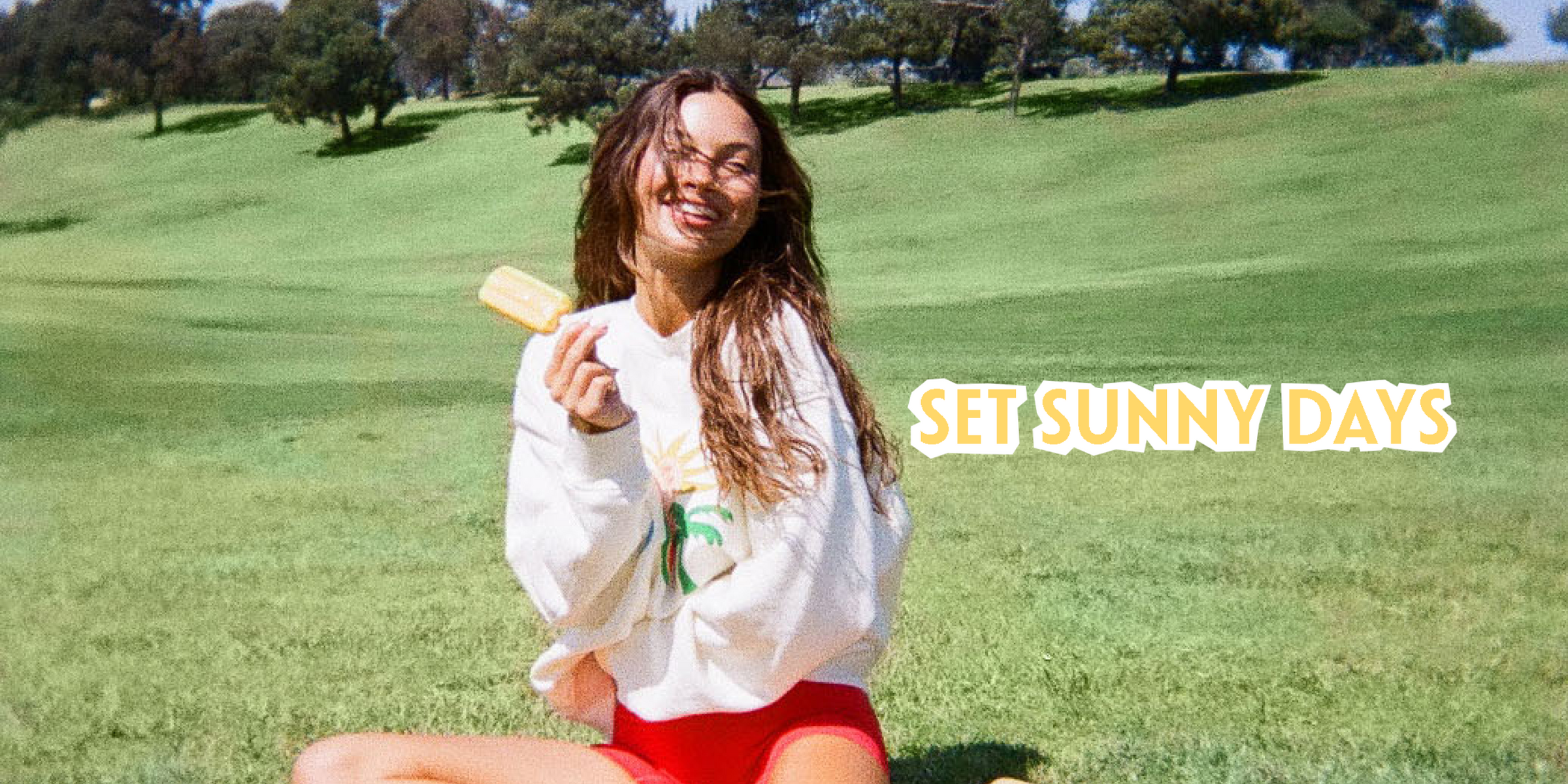 Person sitting on a grassy hill holding a popsicle with 'Set Sunny Days' text.