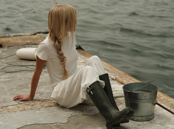 Person in a white dress and black boots sitting on a dock by water