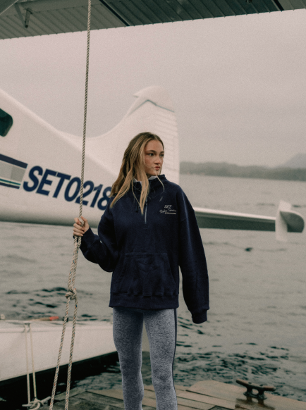 Person wearing a navy blue jacket with a logo, standing on a dock with a boat in the background.