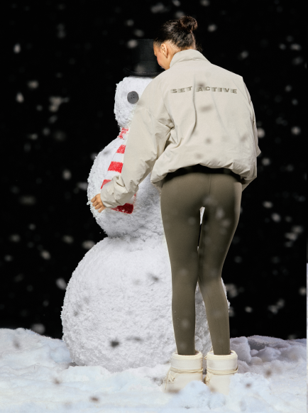 Person in a beige jacket standing next to a snowman in a snowy landscape