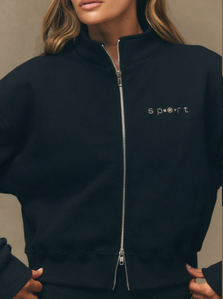 Person wearing a black jacket with 'sport' embroidered on it against a neutral background