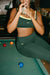 Woman in green sports bra and leggings sitting on a pool table, holding a drink.