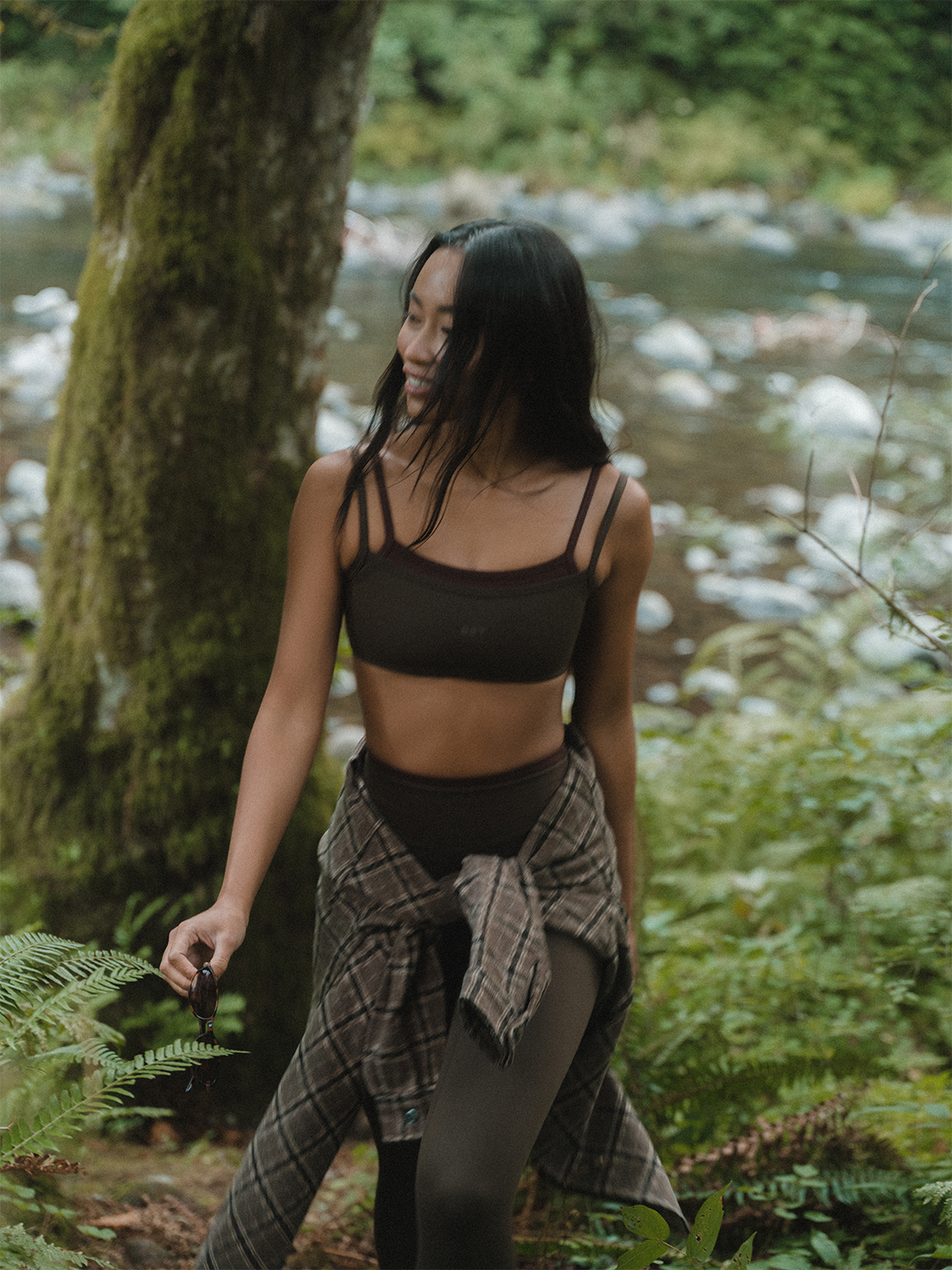 Woman walking through a forest by a stream wearing a black sports bra and plaid skirt.