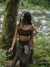 Woman walking through a forest by a stream wearing a black sports bra and plaid skirt.