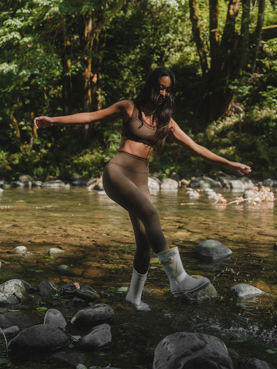 Woman in athletic wear standing in a forest stream with arms outstretched.