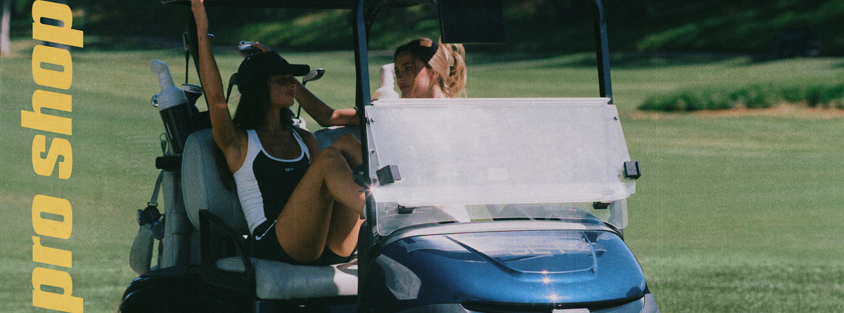 A woman in a black and white athletic outfit sits in a blue golf cart on a grassy golf course, with a blurred background and yellow vertical text reading 'pro shop' along the left side.